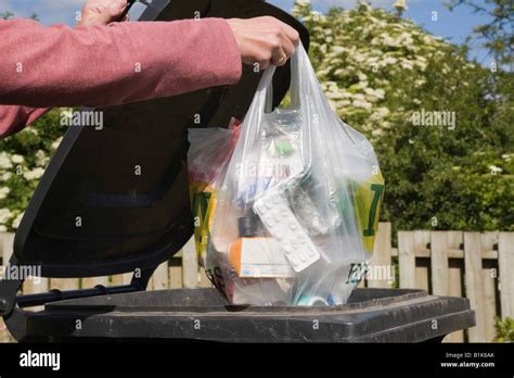 Woman Putting Plastic Bag Of Household Rubbish Into Black Wheelie Bin