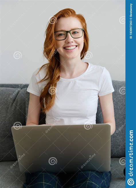 the red haired girl sits on a sofa in a white t shirt and glasses with a laptop smiles broadly