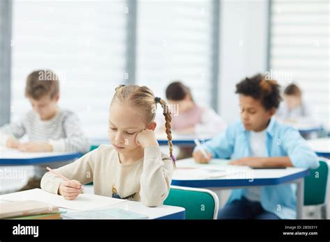 Horizontal Portrait Of Little Blond Girl With Two Plaits Sitting At School Desk Resting Her