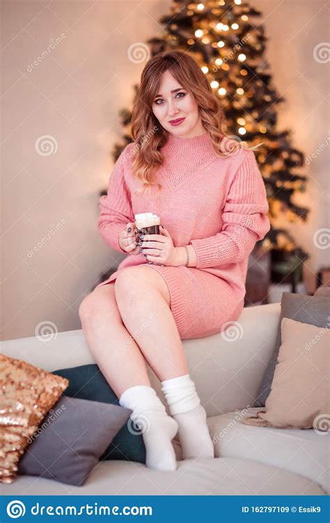 Beautiful Girl In A Pink Sweater And Socks Sits On A Sofa With A Cup Of Hot Drink In Her Hands