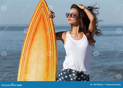 beautiful slender dark haired girl in sunglasses standing near by a yellow surfboard on the