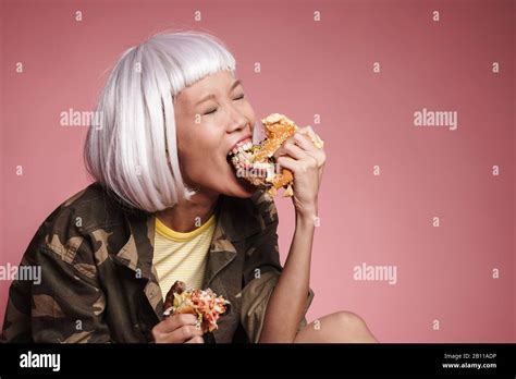 image of wild asian girl wearing white wig eating big burger isolated over pink background stock