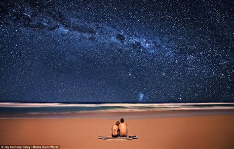 Two People Sitting On A Surfboard In The Sand Under A Night Sky Filled