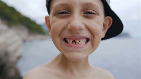 A Cute Toothless Boy In Cap Is Smiling On The Camera At Sea Background