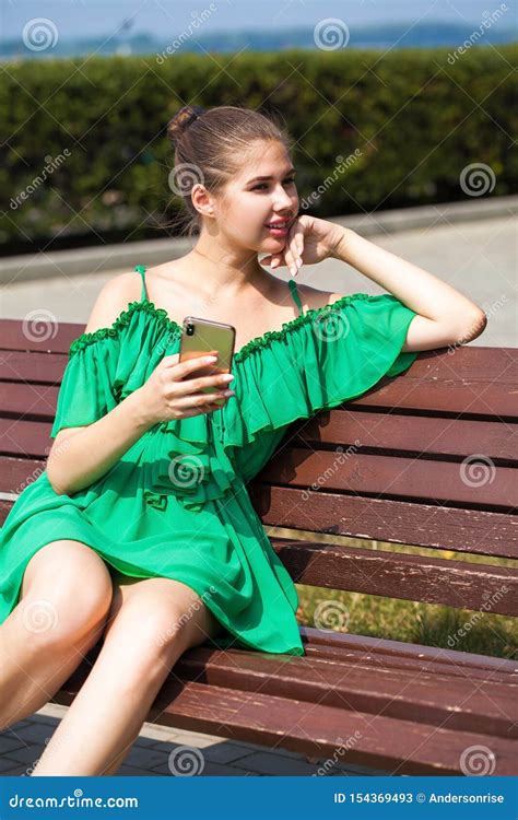 Beautiful Caucasian Girl In Green Dress Sitting On A Bench With A Phone In Hand Stock Image
