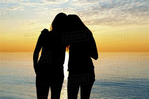 Silhouette Of Two Teenage Girls Standing In An Embrace Looking Out At A Lake At Sunset Woodbine