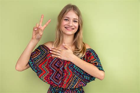 웃는 젊은 금발의 슬라브 소녀가 가슴에 손을 대고 승리의 표시를 하고 있다 무료 사진
