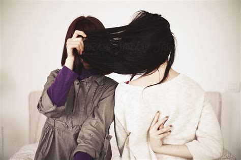 two girls one hiding her face with hair the other seen from the back while sitting on bed by