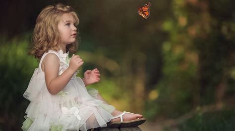 प्यारा छोटी लड़की लकड़ी की बेंच पर बैठी है जो तितली को सफेद पोशाक पहनती