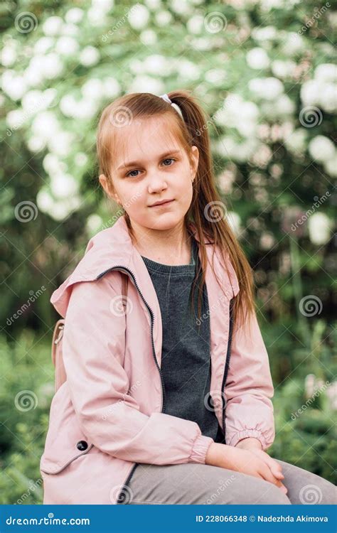 Cute Little Girl Sitting In Park Above Blooming Bush And Looking At
