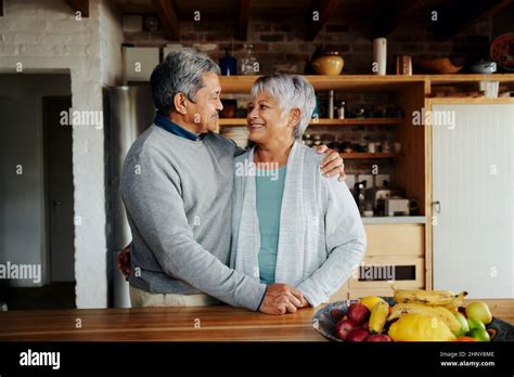 Happily Retired Elderly Biracial Couple Standing Hugging And Smiling At Each Other In Kitchen