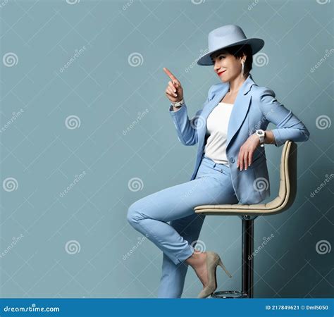 Slyly Smiling Short Haired Brunette Woman In Blue Business Suit And Hat Sits On Stool Pointing