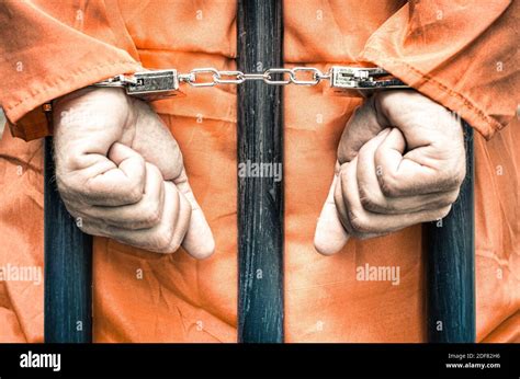 Handcuffed Hands Of A Prisoner Behind The Bars Of A Prison With Orange