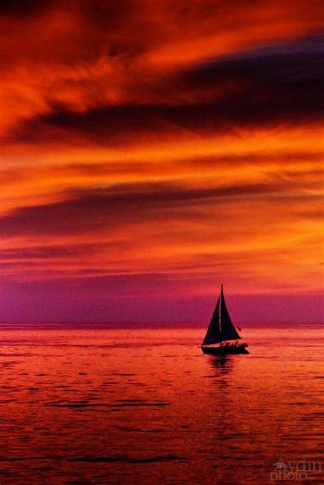 A Sailboat In The Ocean At Sunset With Red And Purple Clouds Above It