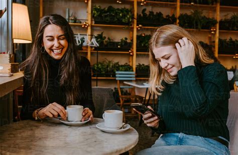 Young Women Laughing At Joke While Drinking Coffee And Using Smartphone During Meeting In Cozy