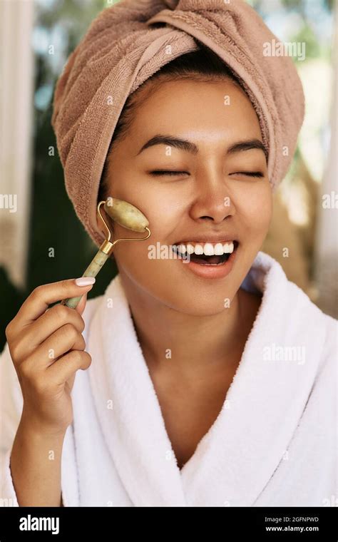 happy asian woman with closed eyes using green jade roller for face massage close up stock