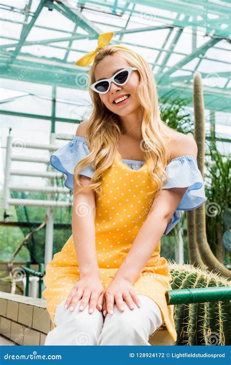 Beautiful Blonde Girl In Sunglasses And Apron Smiling At Camera While Sitting Stock Photo