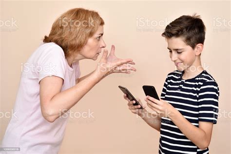 Angry Mother Scolding Her Smiling Son With Two Smartphones Stock Photo