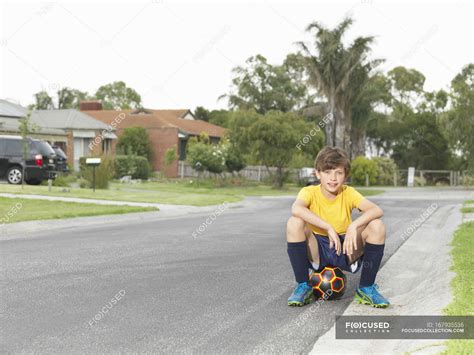 Portrait Of Boy Sitting On Soccer Ball On Suburban Road — Smiling