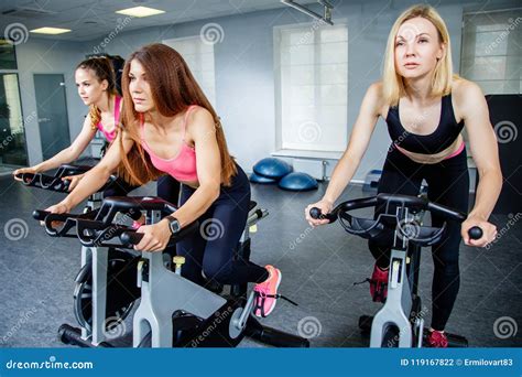 Three Young Women Biking In The Gym Exercising Legs Doing Cardio Workout Cycling Bikes Stock