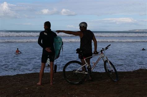 Two Men Standing Next To Each Other On The Beach With A Bike And Surfboard
