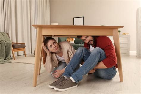 Scared Couple Hiding Under Table In Living Room During Earthquake Stock