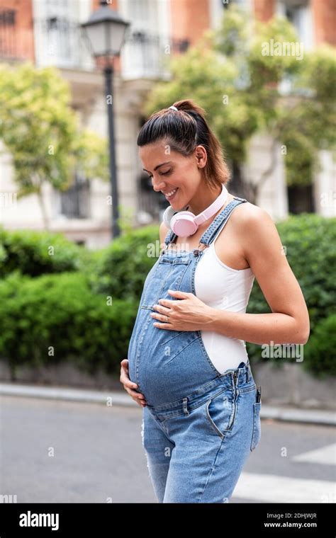 vista lateral de mujer embarazada soñadora en monos de pie la calle mientras disfruta de un