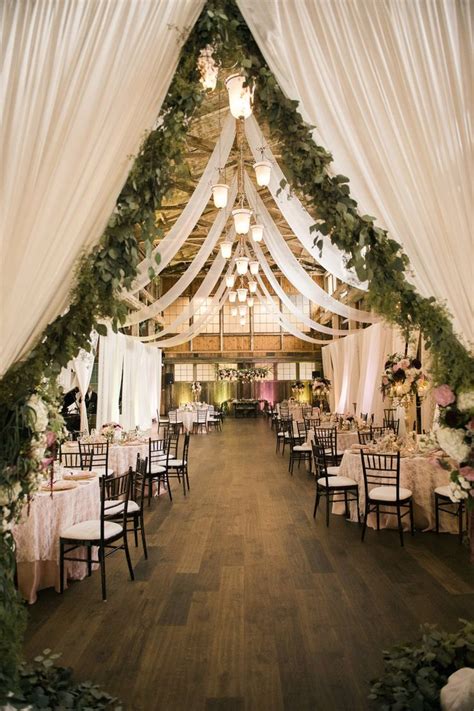 An Indoor Wedding Venue With White Draping And Greenery Draped Over The