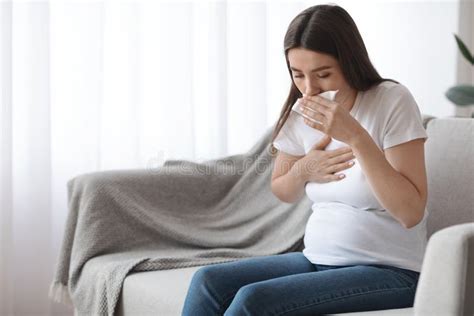 Sick Pregnant Woman Sitting On Couch And Coughing Into Tissue Stock