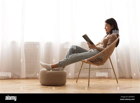 Relaxed Girl Reading Book Sitting In Chair By The Window Stock Photo