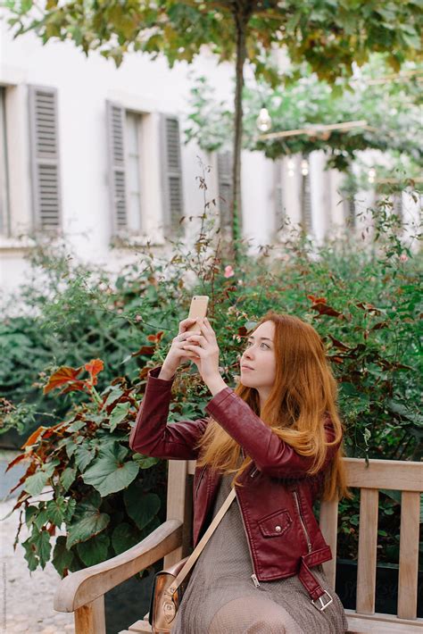 lovely redhead girl sitting on bench in the garden and taking picture with her phone by