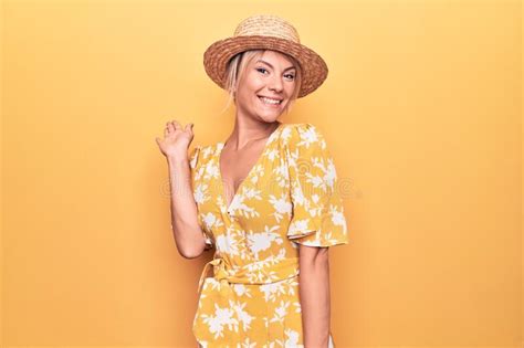 Beautiful Blonde Woman On Vacation Wearing Summer Hat And Dress Over Yellow Background Waiving