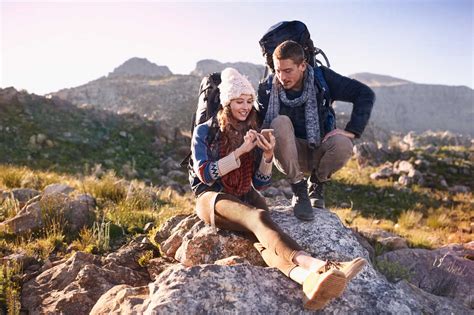 Young Couple With Backpacks Hiking Resting On Rock Using Smart Phone