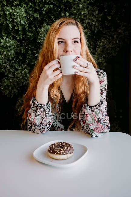 Smiling Redhead Woman With Cup Sitting And Doughnut Sitting Against