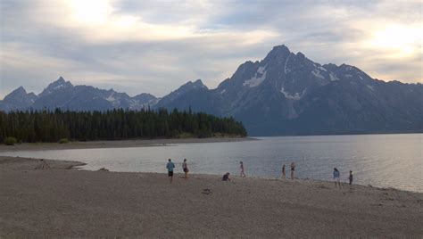 Several People Are Standing On The Shore Of A Lake With Mountains In