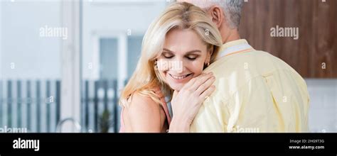Mature Woman Smiling While Hugging Husband In Kitchen Banner Stock