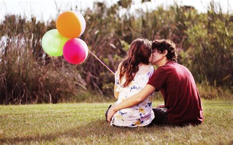 Couple Kissing While Holding Balloons And Sitting On Grass During