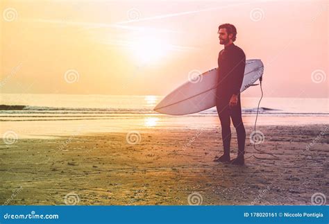 Male Surfer Standing On The Beach Waiting For Waves At Sunset Time