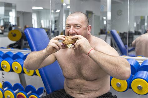 Big Fat Hungry Man Chewing A Hamburger With Meat And Cheese In The Gym