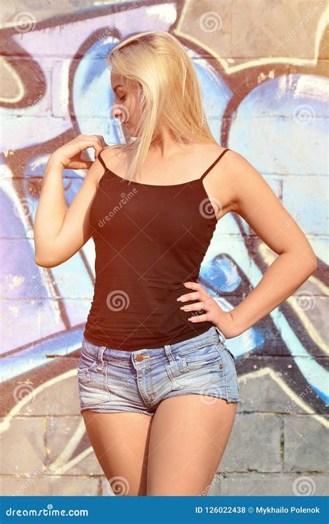 Caucasian Blonde Girl In Denim Shorts And Black Tank Top Posing Against