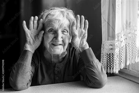 Portrait Of Cheerful Old Woman Holding Wrinkled Hands Near Her Ears Black And White Photo