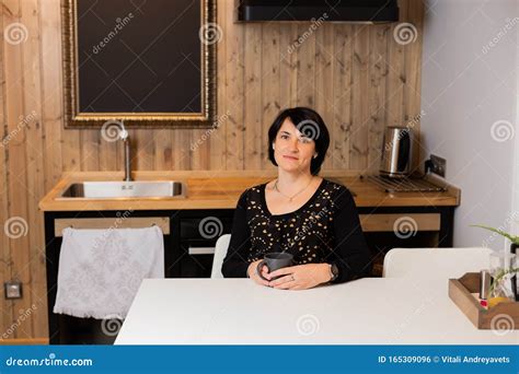 A Beautiful Woman Of 50 Years Old Is Sitting In The Kitchen In A Modern