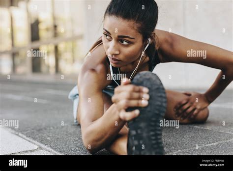 Fitness Woman Stretching Her Leg And Back Sitting Outdoors Listening To Music Wearing Earphones