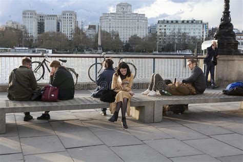 People Sitting On Bench On London S South Bank Overlooking Riv