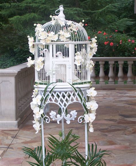 a white birdcage with flowers in it on the ground next to a potted plant