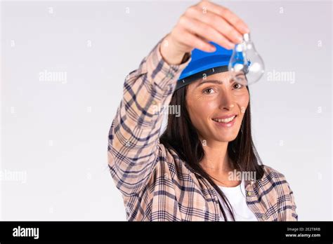 Young Brunette In Hard Hat Posing With Light Bulb And Smiling On White