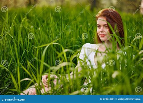 A Pretty Woman Straightens Her Red Hair While Sitting In The Tall Grass In A Light Dress Stock