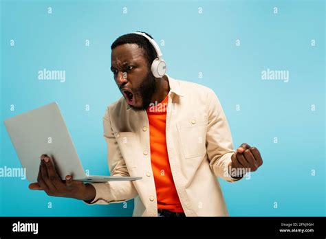 Angry African American Man In Headphones Showing Clenched Fist While Looking At Laptop On Blue