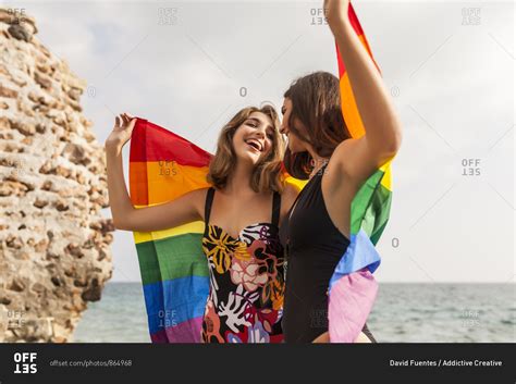 Lesbian Couple Standing On Beach Hugging Wrapped On Colorful Flag Of Lgbt Movement During Summer