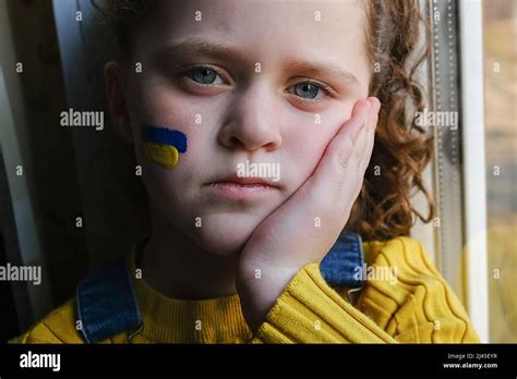 Sad Little Ukrainian Girl With Flag Of Ukraine On Her Face Sitting On Windowsill At Home